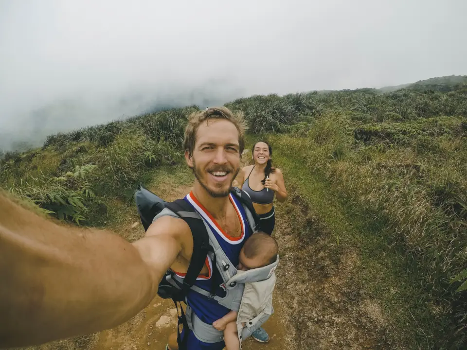 Otec s bábätkom v nosiči si robí selfie na horskom chodníku, za ním mama
